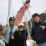 Three adults smile and pose on a sailboat deck among ropes and rigging with sails in the background.
