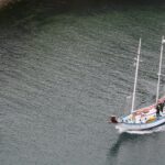 White sailboat with several people wearing life jackets near a rocky shoreline, sailing on dark-green water.