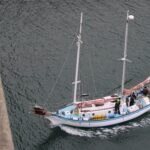 Sailboat with several people near a harbor, viewed from above, white hull with blue trim and orange covers, calm water nearby