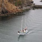 Sailboat with two masts gliding near a rocky coastline along calm water, crew on deck.