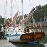 A sailboat named Golden Rule docked at a marina with people on the pier tying it up; tree-lined shore in the background.