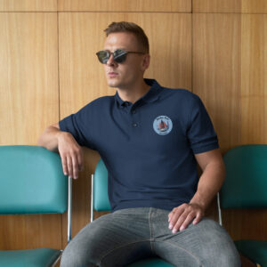 Man wearing round sunglasses and a navy polo with a patch sits against a wood-paneled wall, in front of teal chairs.